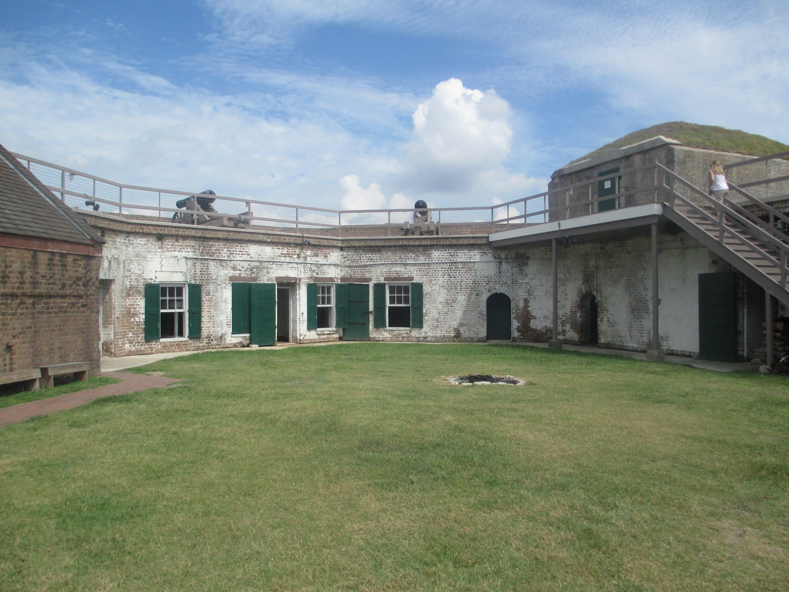 A historic brick fort, Old Fort Jackson Savannah GA, with green doors and windows surrounds a grassy courtyard. Cannons sit atop the walls, and a wooden staircase leads to an upper level beneath a partly cloudy sky.