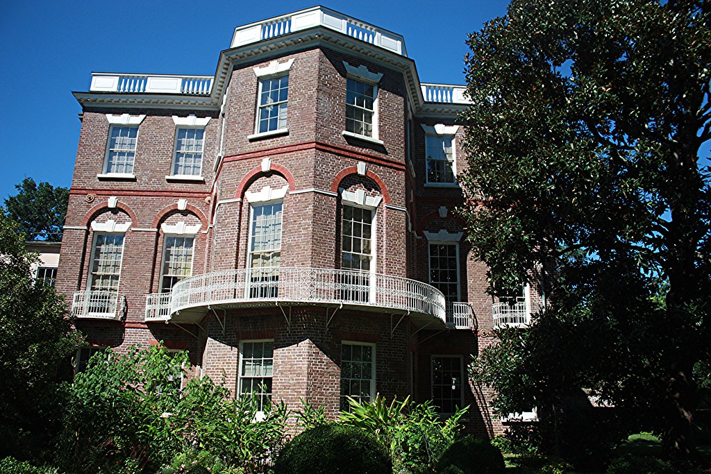 A large historic brick building with tall windows, white balcony railings, and decorative trim—one of the notable historic landmarks in Charleston SC—surrounded by lush greenery and trees under a clear blue sky.