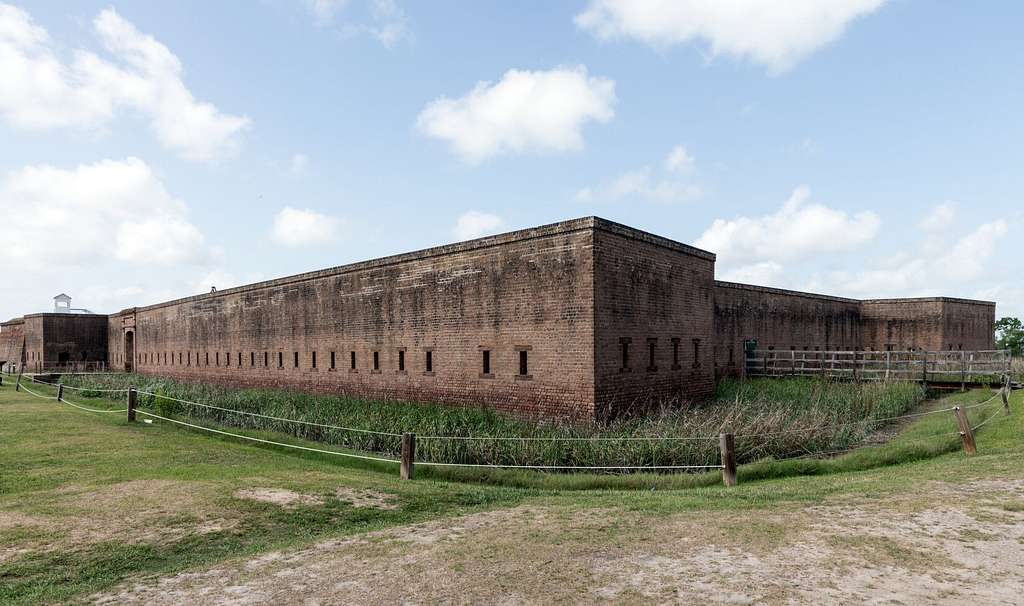 A large, rectangular, historic brick fort—Old Fort Jackson Savannah GA—with small windows, surrounded by grassy grounds, a rope fence, and a wooden walkway under a partly cloudy sky.