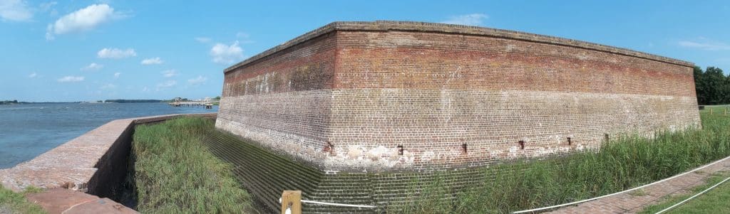 A large brick fortification with thick walls sits next to a body of water under a blue sky, surrounded by grass and a white rope barrier—this is Old Fort Jackson in Savannah, GA.