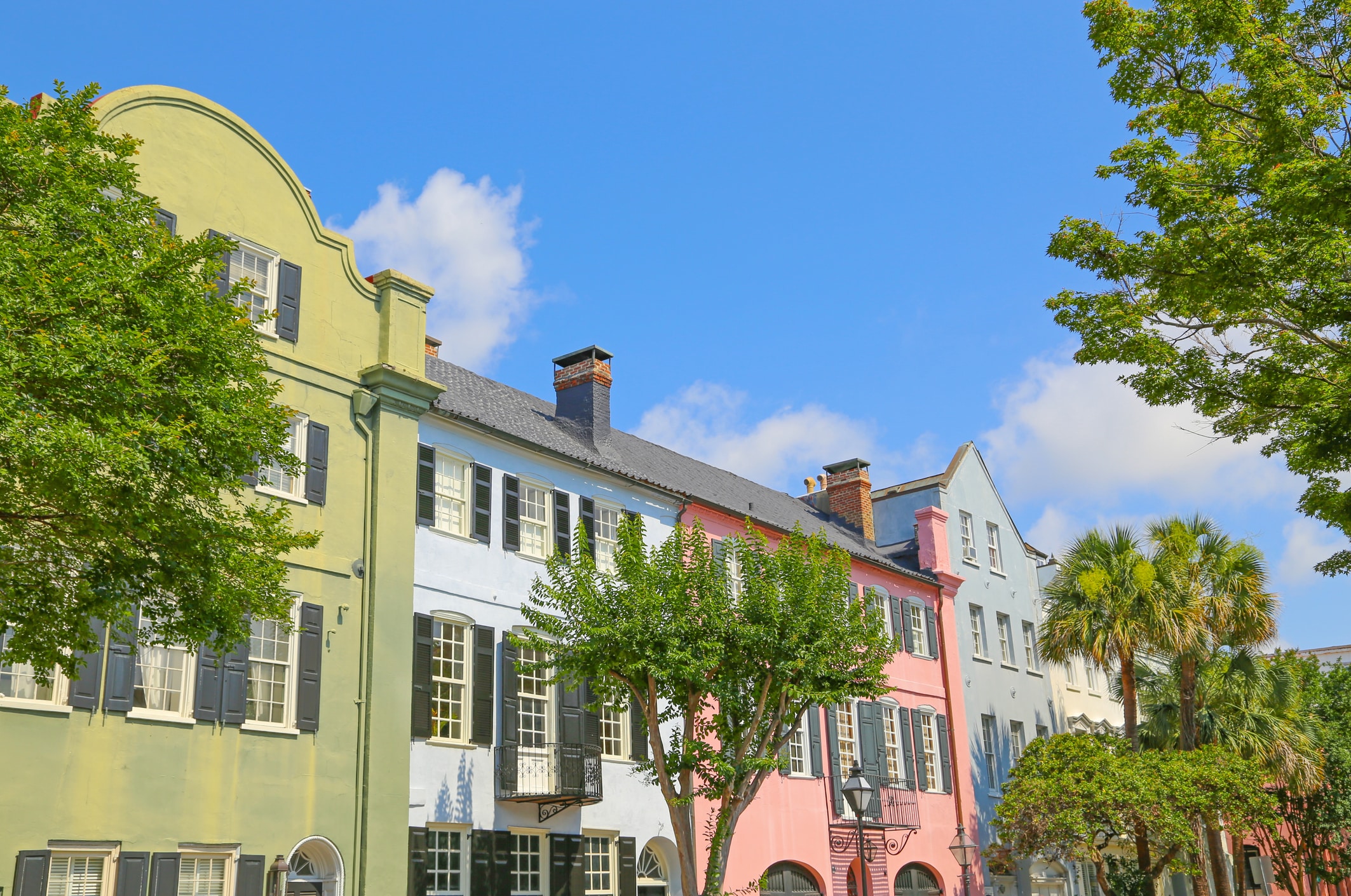 Colorful historic row houses, some of the most charming historic landmarks in Charleston SC, with green, blue, and pink facades stand under a bright blue sky with scattered clouds, framed by leafy trees in the foreground.