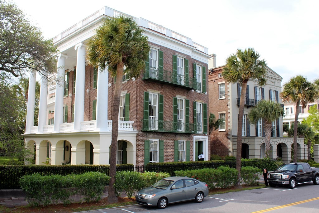 A large, historic brick house—one of the notable historic landmarks in Charleston SC—with tall white columns and green shutters sits behind palm trees and a hedge. Two cars are parked on the street in front of the house on a sunny day.
