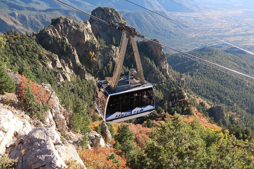 A cable car labeled "Sandia Peak" travels above a forested mountain landscape, with rocky cliffs and a broad valley visible in the background—an ideal scene for a New Mexico Field Trip adventure.