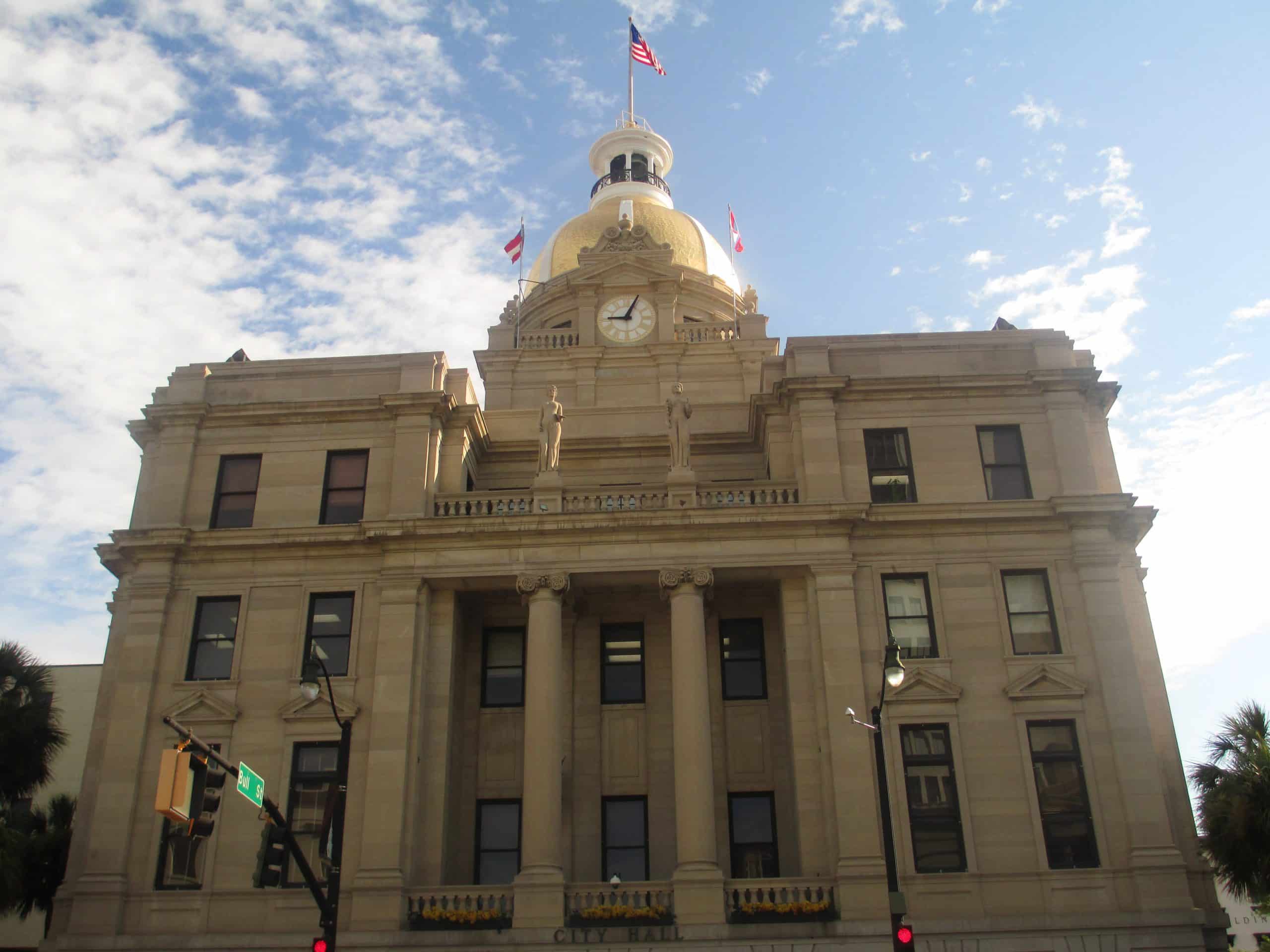 A historic stone building with columns, arched windows, and a large golden dome topped with an American flag stands under a partly cloudy sky. A sign at the entrance reads “City Hall”—a classic stop on any field trip in Savannah, GA.