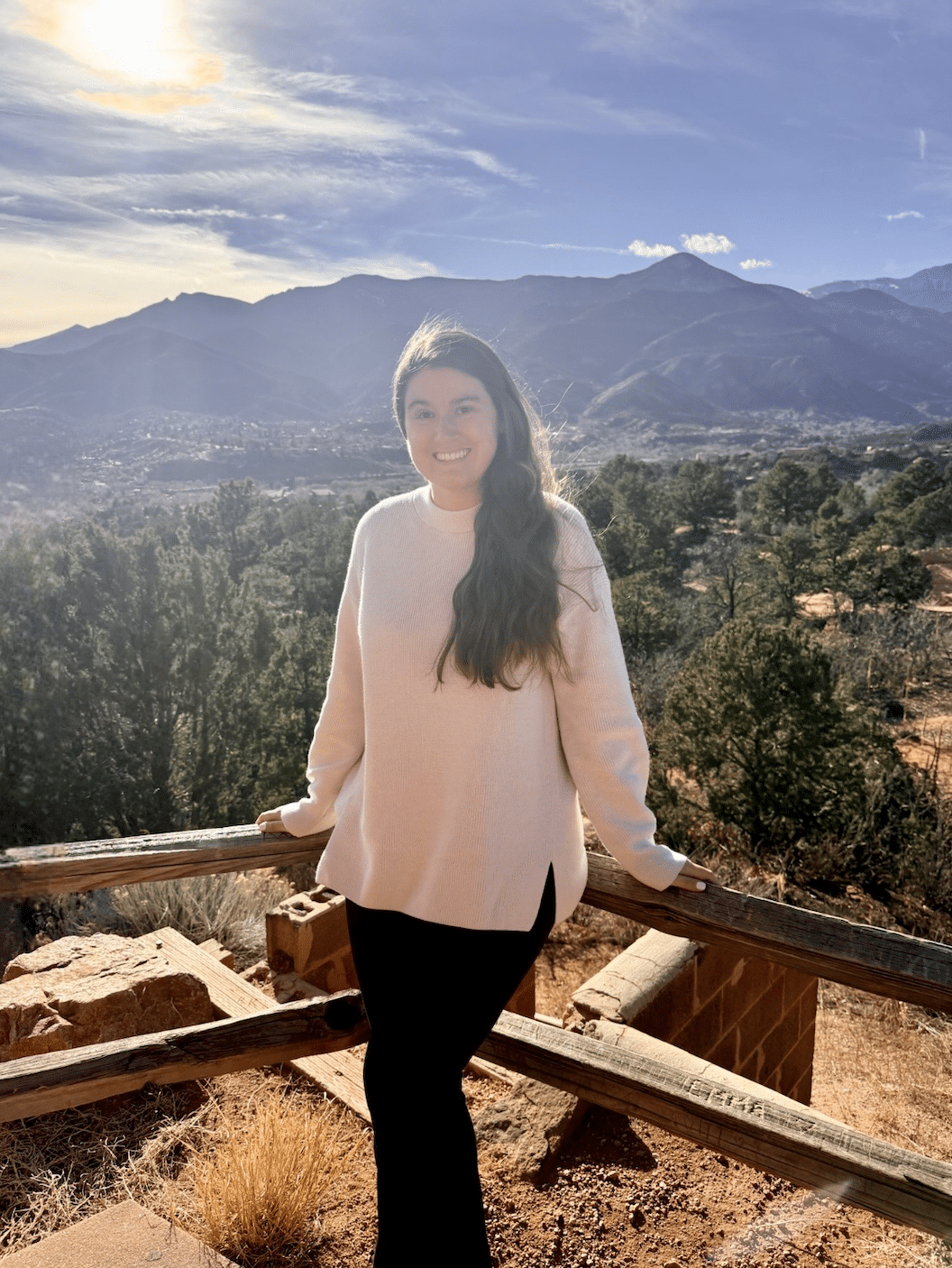 A woman in a white sweater and black pants stands smiling in front of a wooden fence, with mountains, trees, and a bright sun in the background under a blue sky, capturing all that’s beautiful about the outdoors.