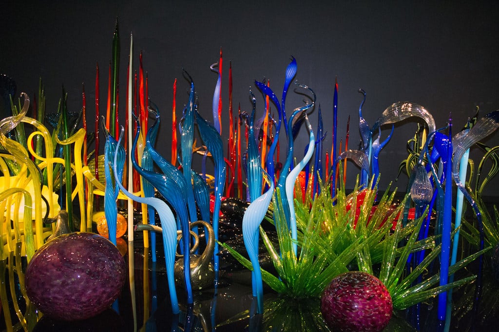 Colorful glass sculptures resembling tall reeds, twisted shapes, spiky grass, and large marbled orbs are arranged against a dark background, evoking the vibrant artistry often admired on a Seattle School Trip.