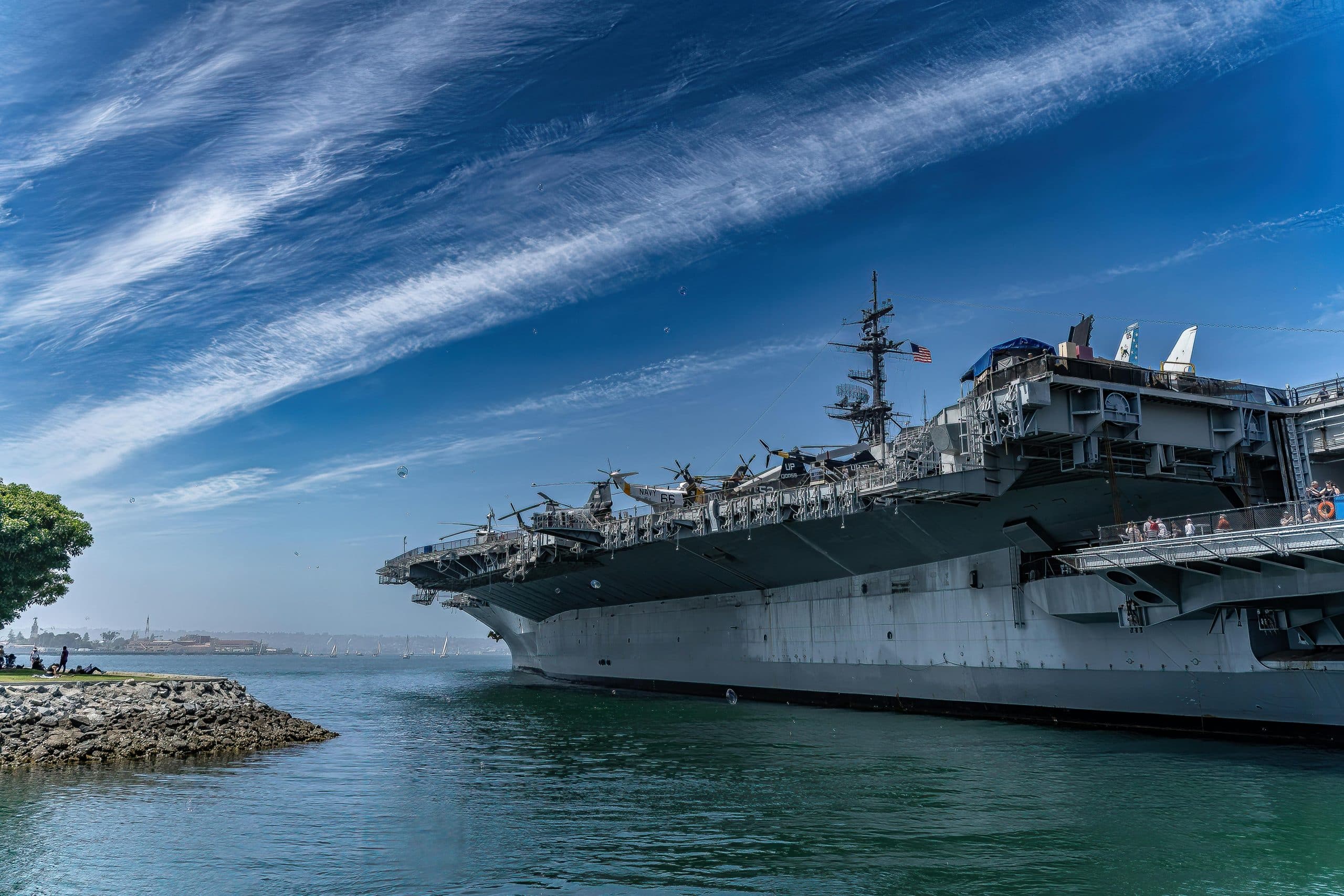 A large aircraft carrier is docked in calm waters under a blue sky with streaks of clouds. People along the waterfront on the left enjoy scenic views, making it one of the best educational tours in California. Trees and a distant shoreline complete the scene.