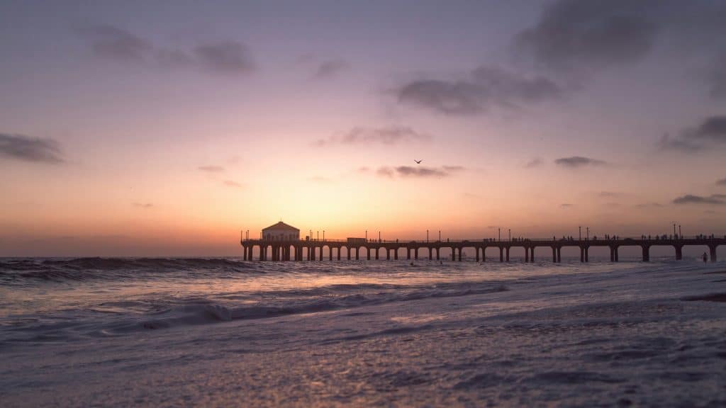 A long pier stretches into the ocean at sunset, with gentle waves in the foreground and a pastel sky overhead. On this Los Angeles field trip, silhouetted people walk along the pier, and a small building sits at the far end.
