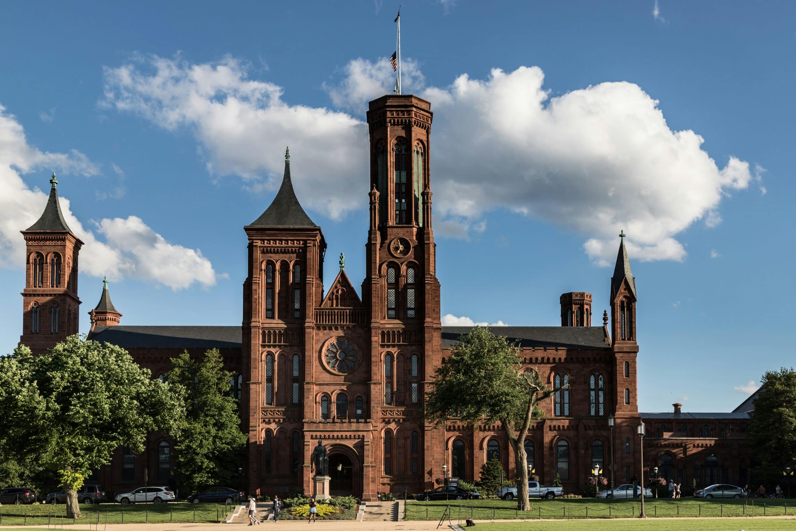A large red-brick building with tall towers and arched windows stands under a blue sky with scattered clouds; trees and people are visible in front—an iconic scene often found in the nation's capital, where DC stands for District of Columbia.
