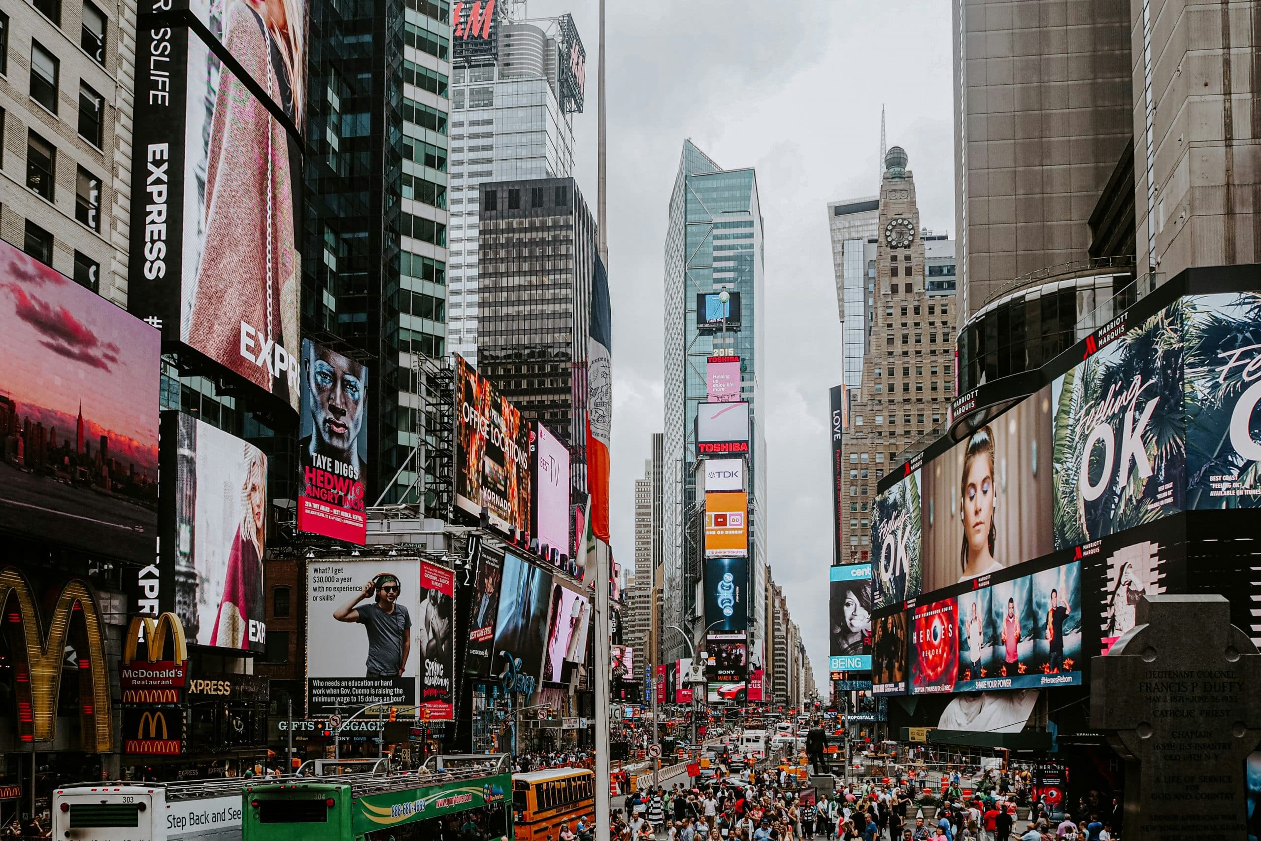 A bustling Times Square scene, perfect for School Field Trips to NYC, with crowds of people, tall buildings covered in bright digital billboards and advertisements, city traffic, and a cloudy sky overhead.