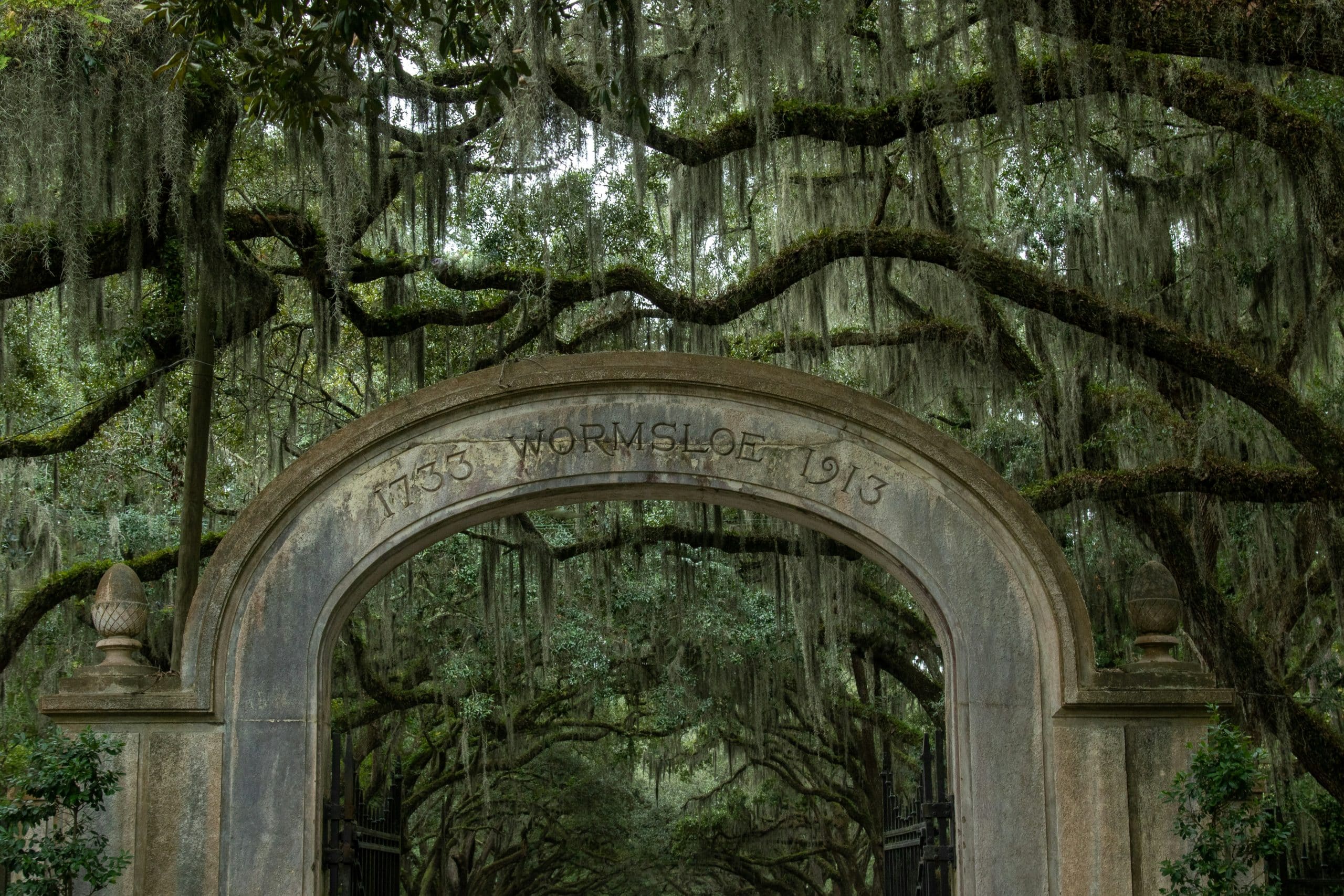 A stone archway with “1733 Wormsloe 1913” engraved on it stands beneath large oak trees draped in Spanish moss at the entrance to Wormsloe Historic Site, a popular Field Trip Savannah GA destination.