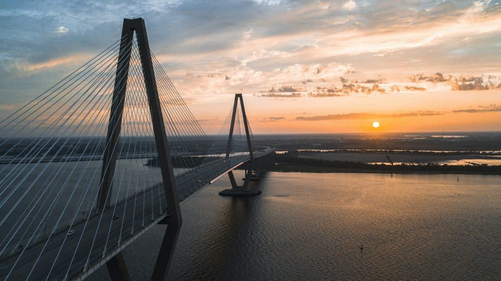 A cable-stayed bridge spans across a wide river at sunset, with orange and yellow sunlight reflecting on the water—an ideal scene for Charleston Field Trips, where students can enjoy breathtaking views and learn about local landmarks.