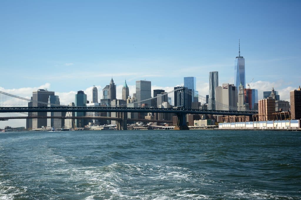 A view of the Manhattan skyline with skyscrapers and One World Trade Center, seen across the East River with the Manhattan Bridge in the foreground under a clear blue sky—a perfect scene for School Field Trips to NYC.