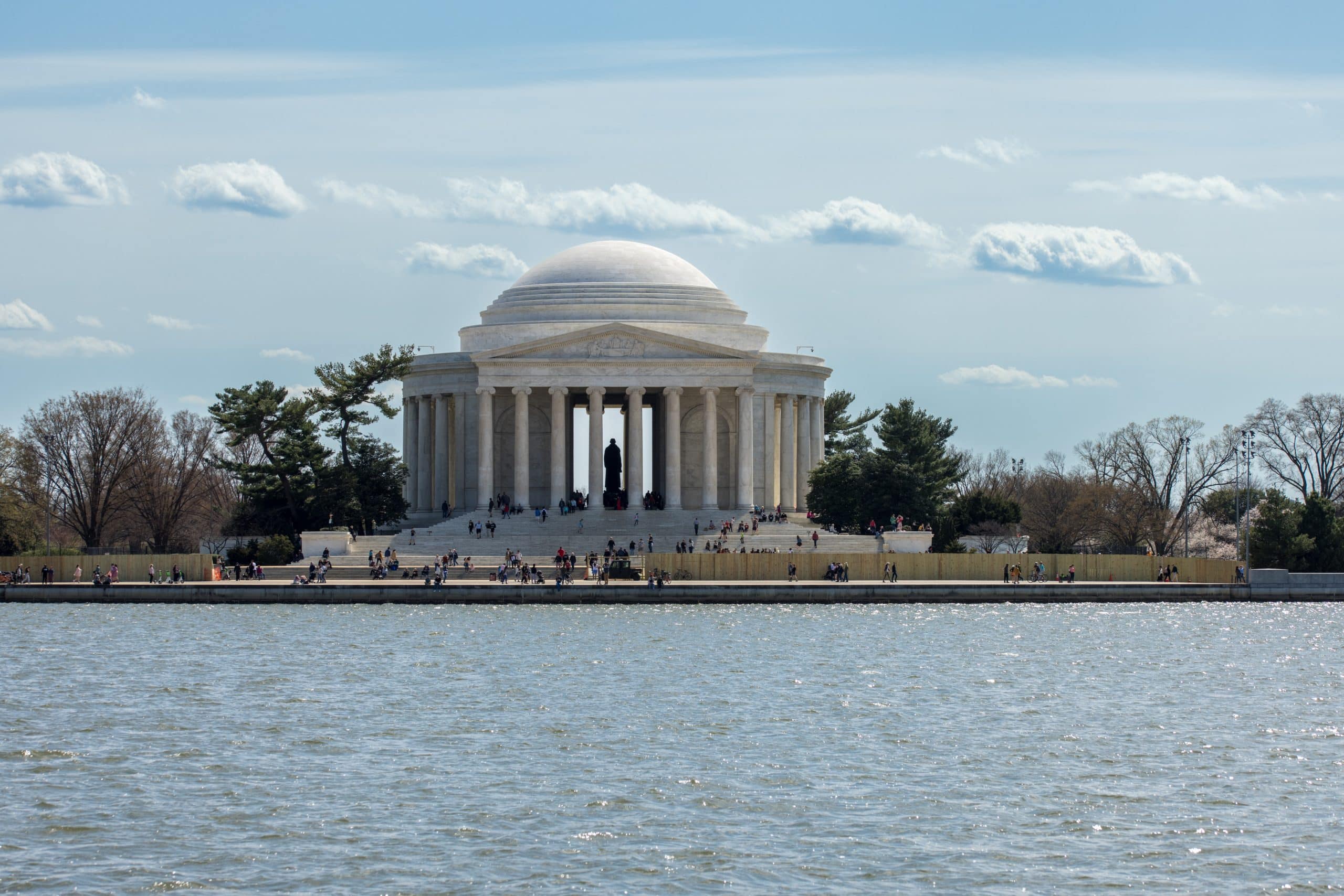 The Jefferson Memorial stands beside the Tidal Basin in Washington, D.C.—short for District of Columbia—with people walking and sitting along the waterfront under a partly cloudy sky.