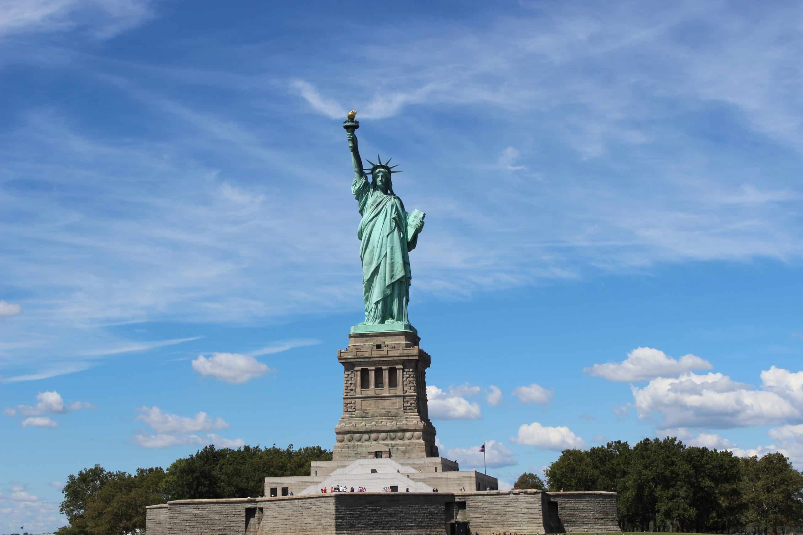 The Statue of Liberty stands tall against a blue sky with scattered clouds, surrounded by greenery at its base on Liberty Island—a must-see landmark for school field trips to NYC.