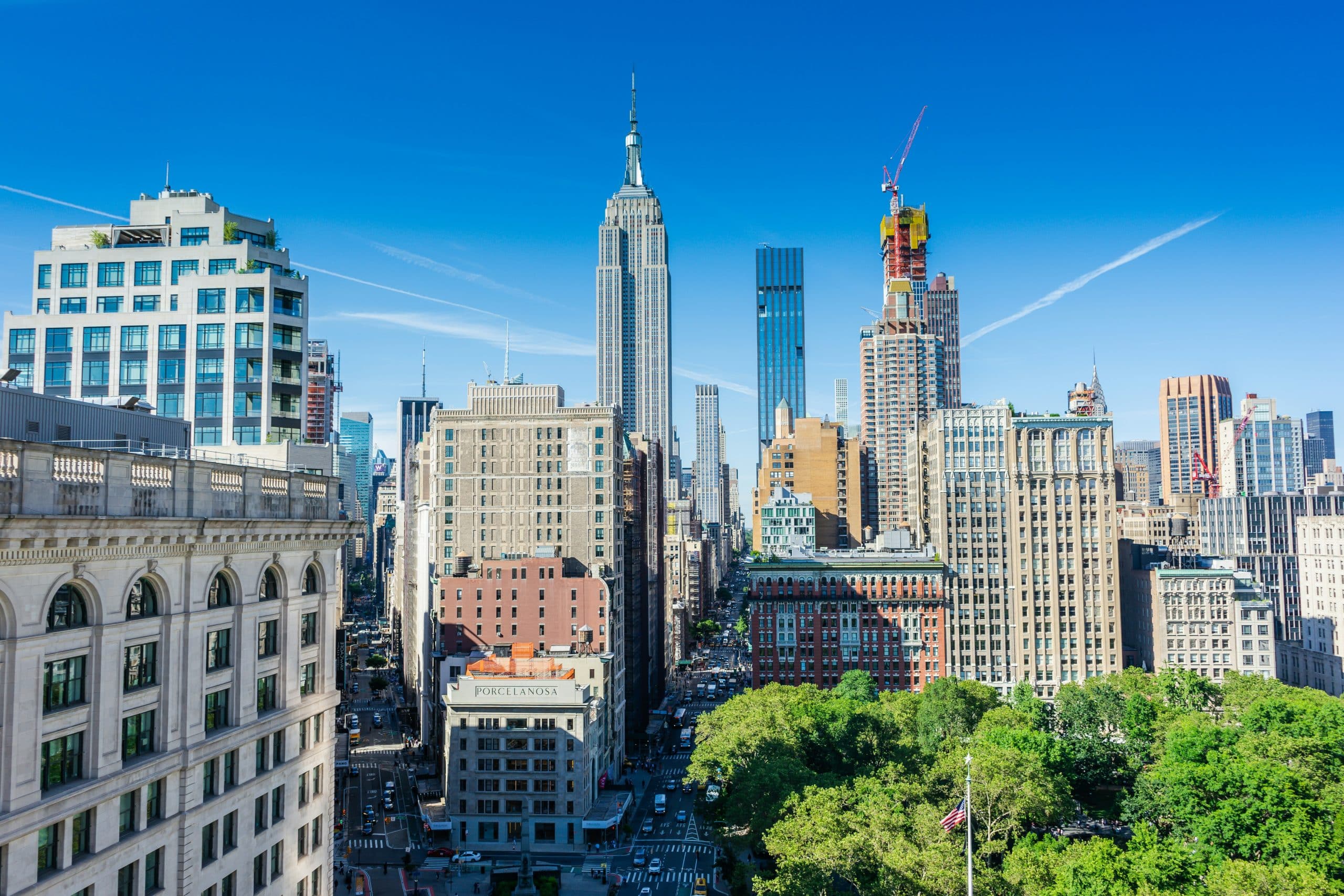 A sunny cityscape of Midtown Manhattan, New York City, with the Empire State Building and other skyscrapers, invites you to start exploring American history through NYC landmarks, with streets below and green trees in the foreground under a clear blue sky.