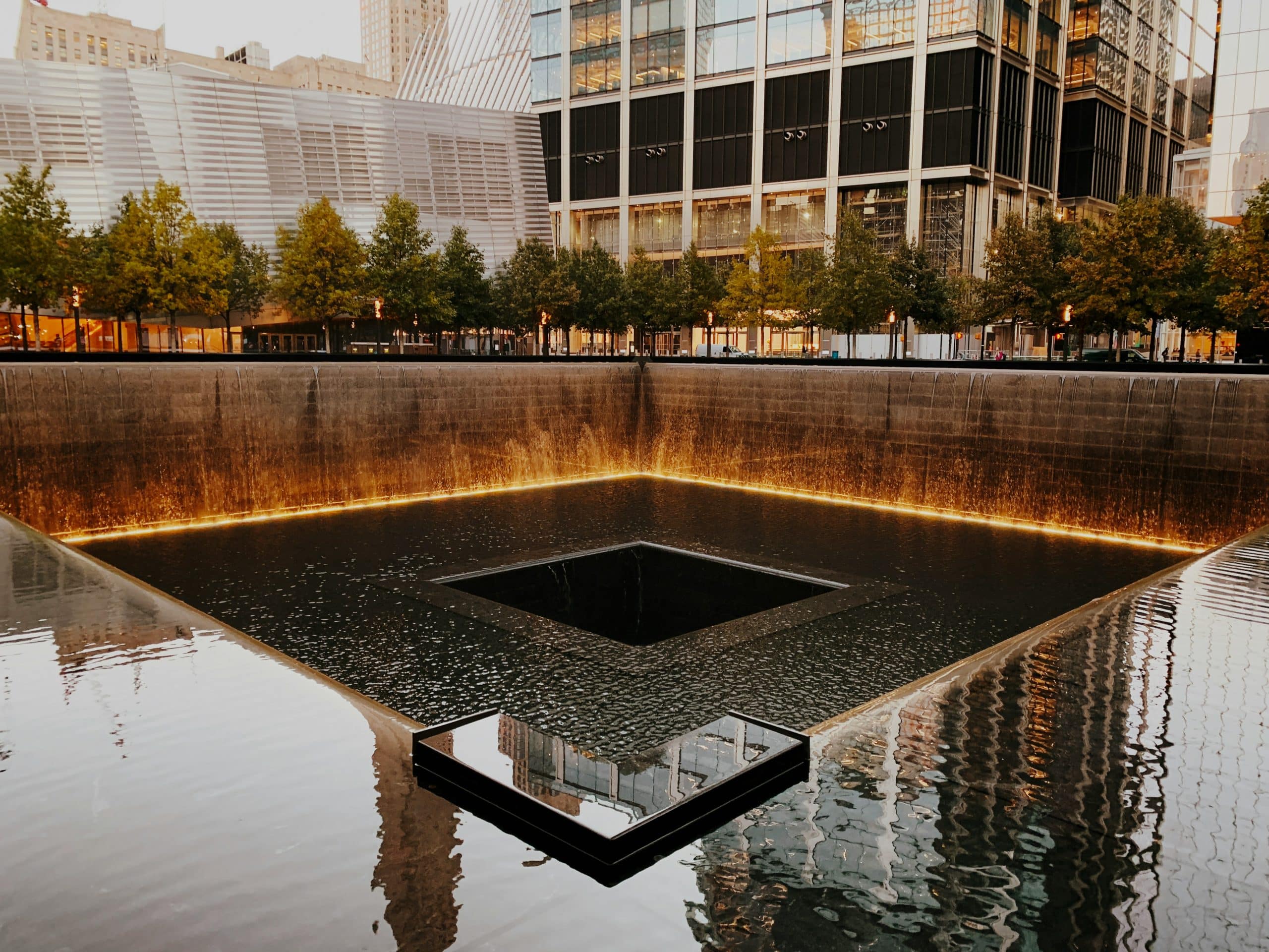 A large square reflecting pool with a central void, surrounded by cascading water and illuminated edges at the 9/11 Memorial in New York City—a moving site often visited during school field trips to NYC, with trees and modern buildings in the background.