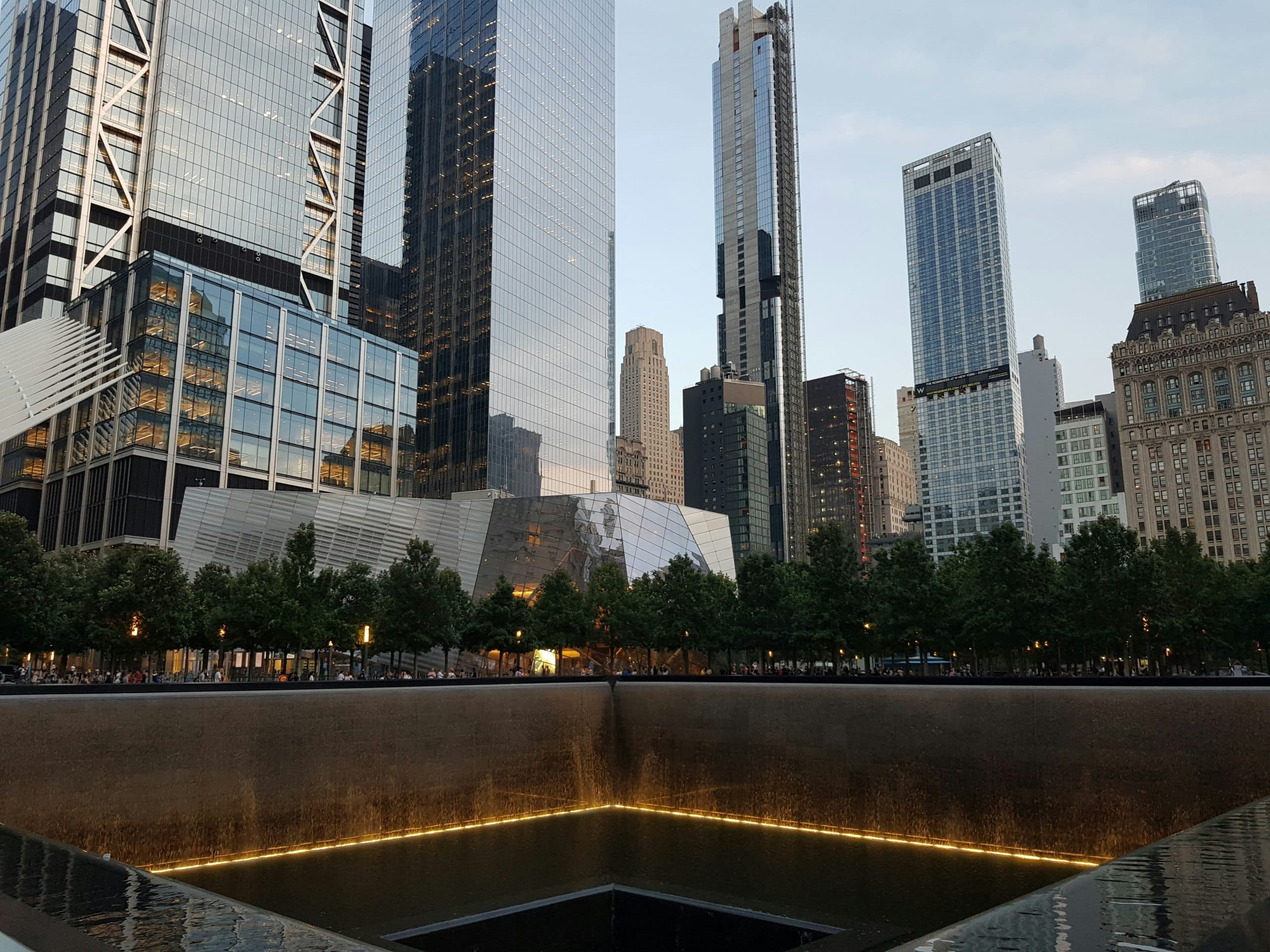 The image shows the 9/11 Memorial reflecting pool in New York City, surrounded by trees and modern skyscrapers at dusk, offering a poignant moment for those exploring American history through NYC landmarks.