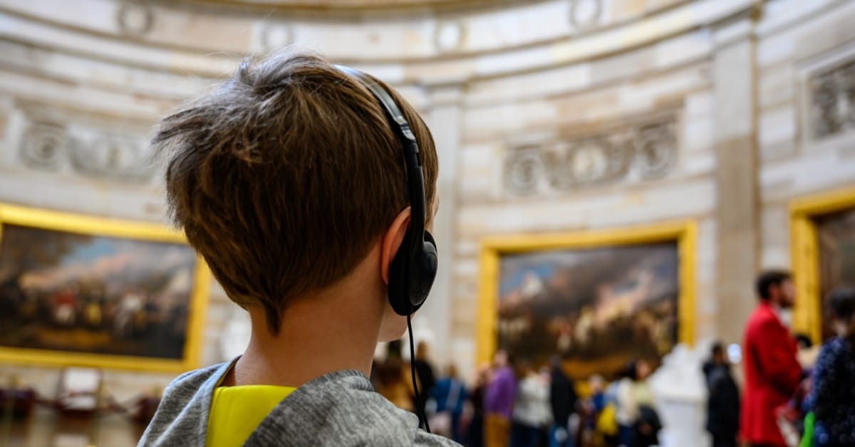 A young boy stands with a headset on in the capital building. He's on a tour with his class and other people in the room.