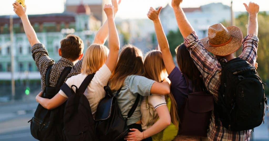 A group of junior high students stand together with their backpacks on a school trip. They have their arms up with joy.