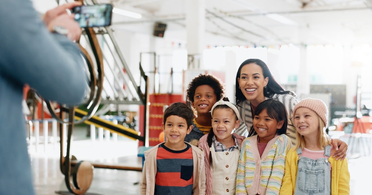 A group of young students with their teacher pose for a photo on their school trip. Someone takes their photo.