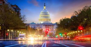 A nighttime view of the capitol building in Washington, DC. The street lights are red and cars are driving.