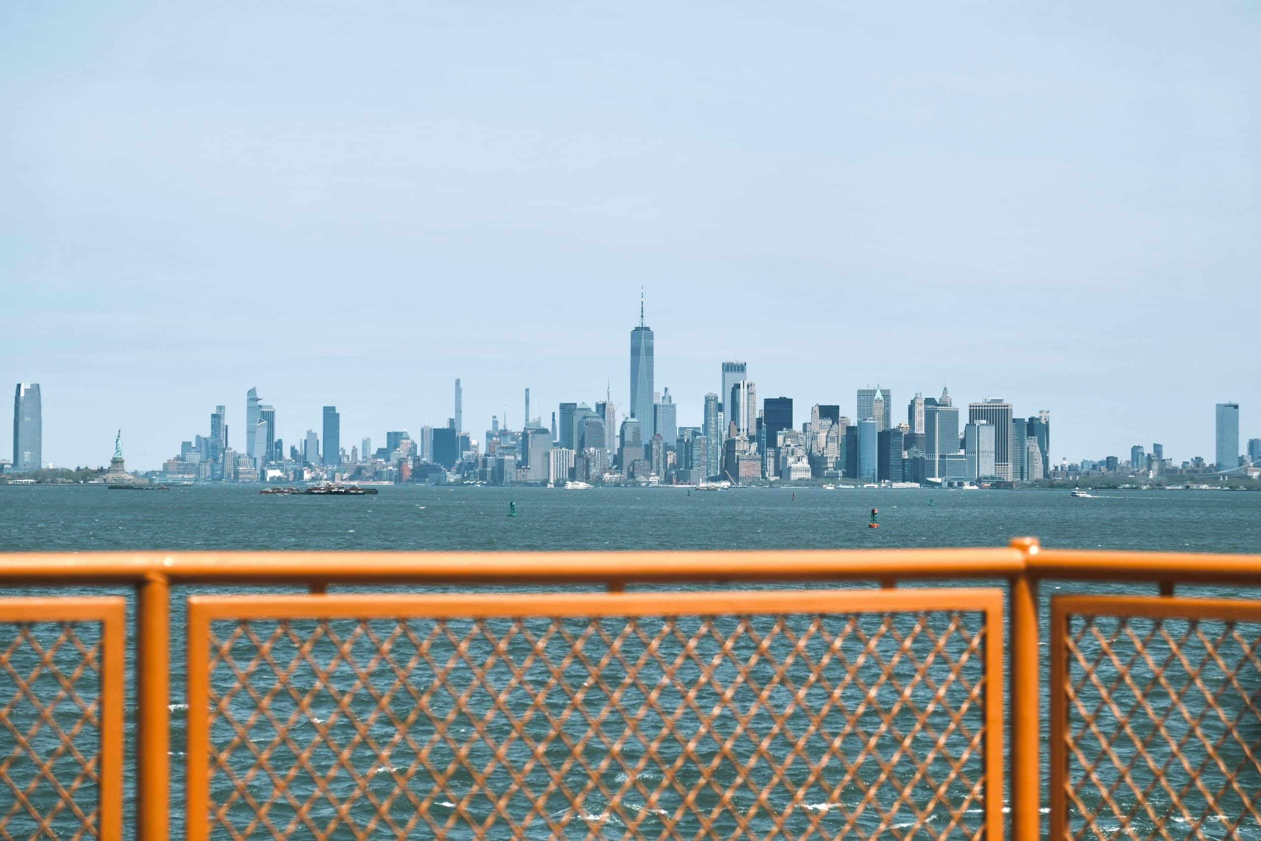 A clear view of the New York City skyline with skyscrapers, seen across the water from behind the orange railing of a Staten Island Ferry—perfect for School Field Trips to NYC.