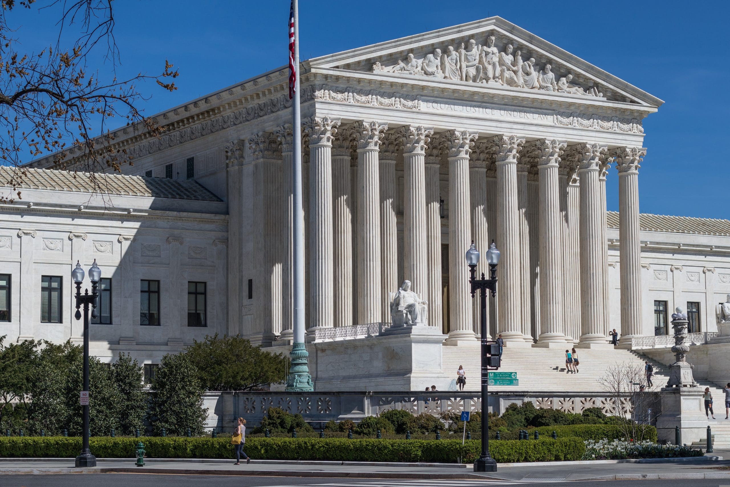 The United States Supreme Court building with tall columns, statues, and people walking up the steps in Washington DC—where DC stands for District of Columbia; an American flag waves under a clear blue sky.