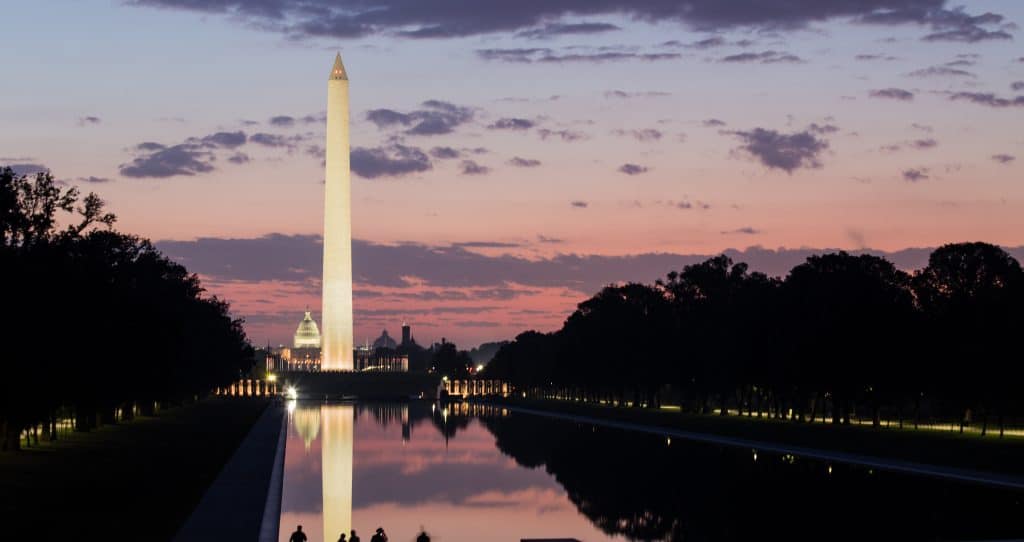 The Washington Monument illuminated at dusk reflects in the Lincoln Memorial Reflecting Pool, with the U.S. Capitol in the background and a pink and purple sky overhead—capturing the essence of what “DC” stands for in Washington DC.
