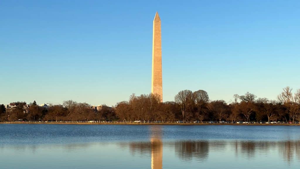 The Washington Monument stands tall against a clear blue sky, reflected in the calm waters of the Tidal Basin—a highlight for any 8th Grade Washington DC Trip Itinerary, with bare trees and distant buildings lining the shoreline.