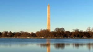 The Washington Monument stands tall against a clear blue sky, reflected in the calm waters of the Tidal Basin—a highlight for any 8th Grade Washington DC Trip Itinerary, with bare trees and distant buildings lining the shoreline.