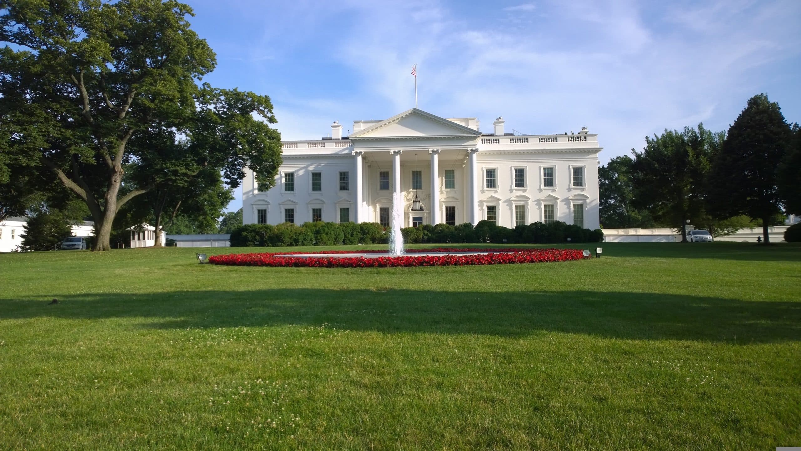 The White House in Washington, D.C.—where “D.C.” stands for District of Columbia—features a fountain and a circular bed of red flowers on its lush green lawn, framed by trees under a clear blue sky.