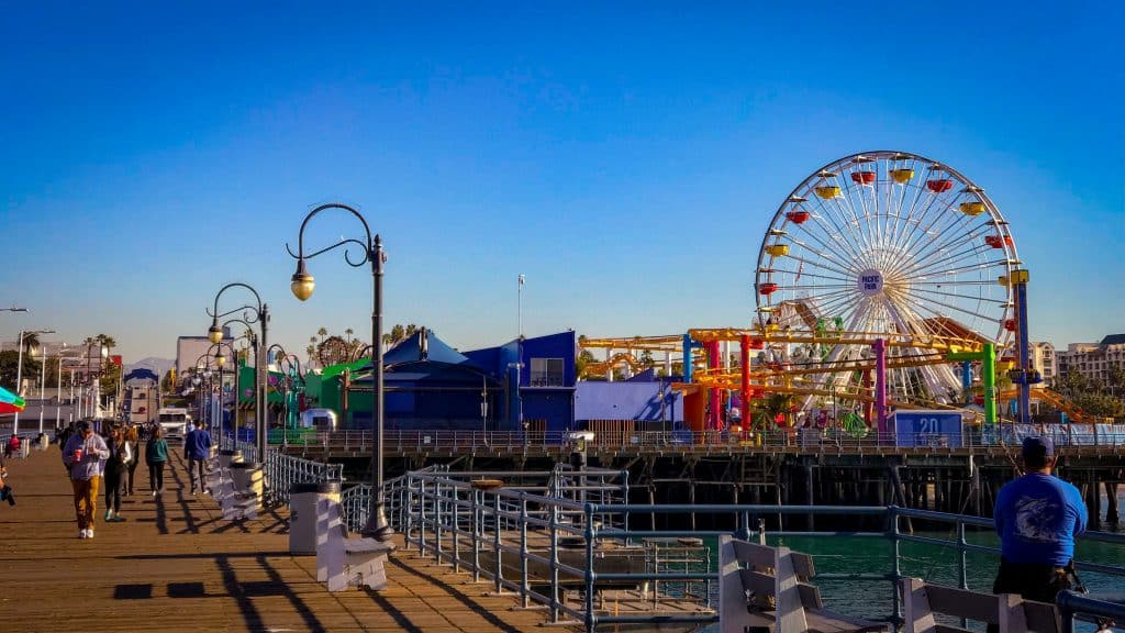 A sunny day at the pier with people walking, colorful buildings, a large Ferris wheel, and street lamps under a clear blue sky—perfect for a Los Angeles school trip by the ocean next to the wooden boardwalk.
