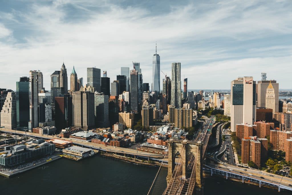 Aerial view of the New York City skyline with tall skyscrapers, the Brooklyn Bridge in the foreground, and the Hudson River on the left—perfect for exploring American history through NYC landmarks under a partly cloudy sky.