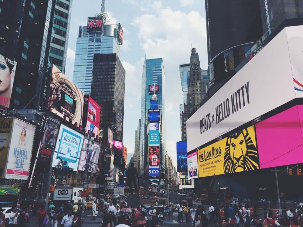 A busy Times Square in New York City with large digital billboards, including ads for “Hello Kitty” and “The Lion King,” surrounded by tall buildings and crowds—an iconic spot for exploring American history through NYC landmarks.