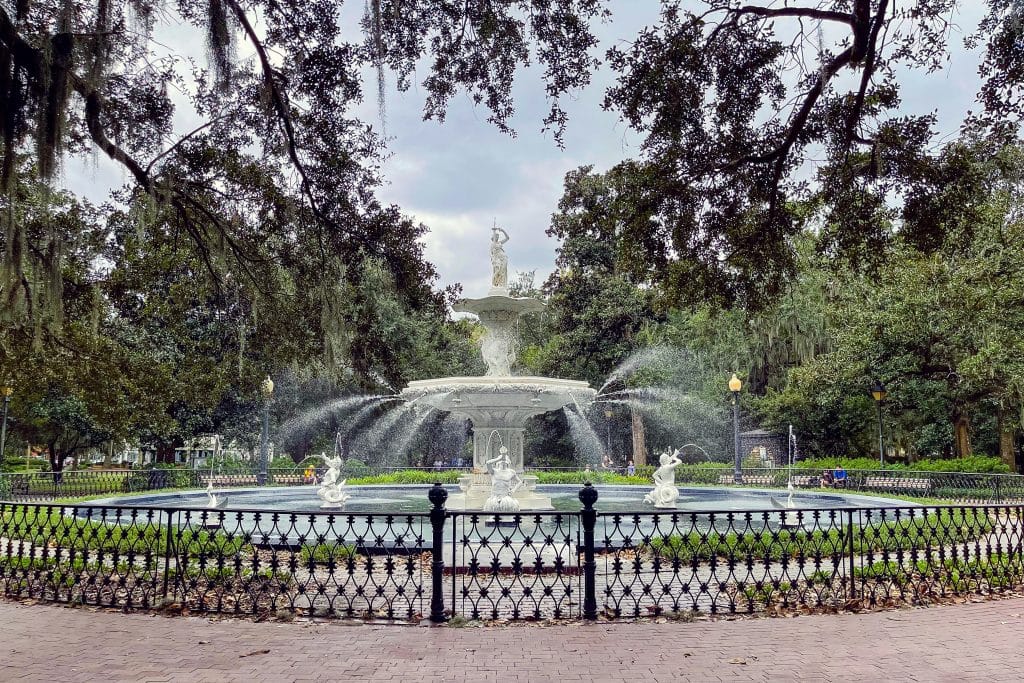A large ornate fountain with multiple water streams stands in the center of a park, surrounded by trees, greenery, and a black iron fence—perfect for a Field Trip in Savannah GA under a cloudy sky.