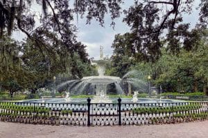 A large ornate fountain with multiple water streams stands in the center of a park, surrounded by trees, greenery, and a black iron fence—perfect for a Field Trip in Savannah GA under a cloudy sky.