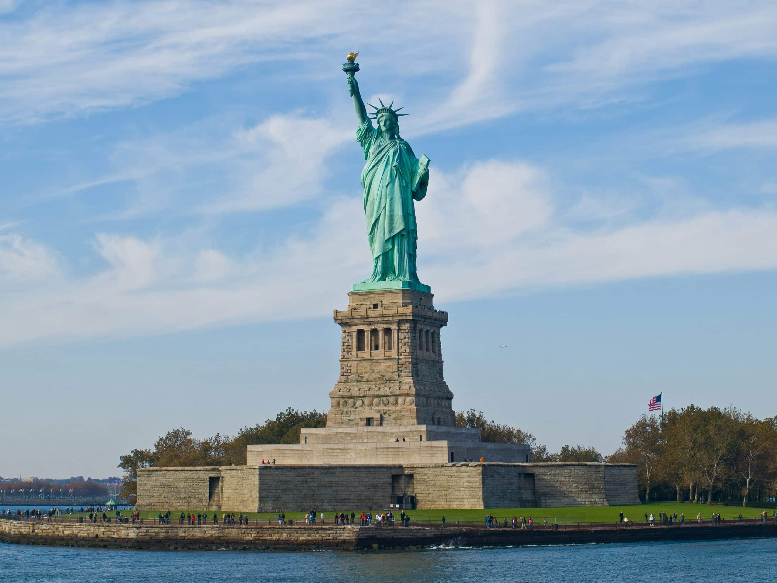 The Statue of Liberty stands on Liberty Island with blue sky and wispy clouds overhead. People gather around the base, an American flag flies to the right, and water surrounds the island—an iconic stop when exploring American history through NYC landmarks.