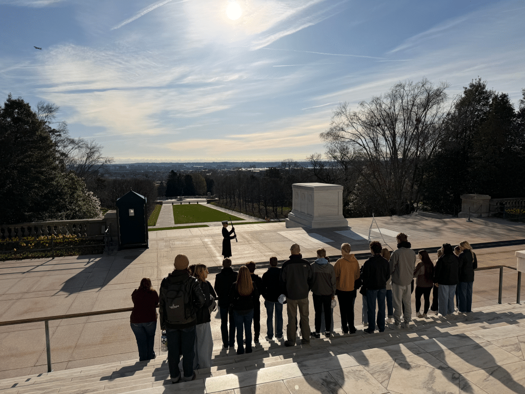 A group of people watches a guard walk near the Tomb of the Unknown Soldier at Arlington National Cemetery on a sunny day, with trees and a distant city skyline in the background.