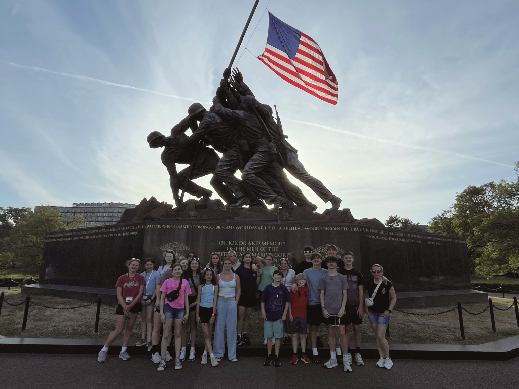 A group of students poses in front of the U.S. Marine Corps War Memorial (Iwo Jima Memorial) in Arlington, VA, featuring a large bronze sculpture of Marines raising the American flag.