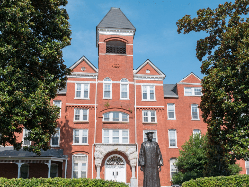 A large red-brick building with a central tower and arched windows, surrounded by trees. In front stands a statue of a person in academic robes. The entrance reads “GRANT HALL,” a site for Landmark Educational Tours Registration.