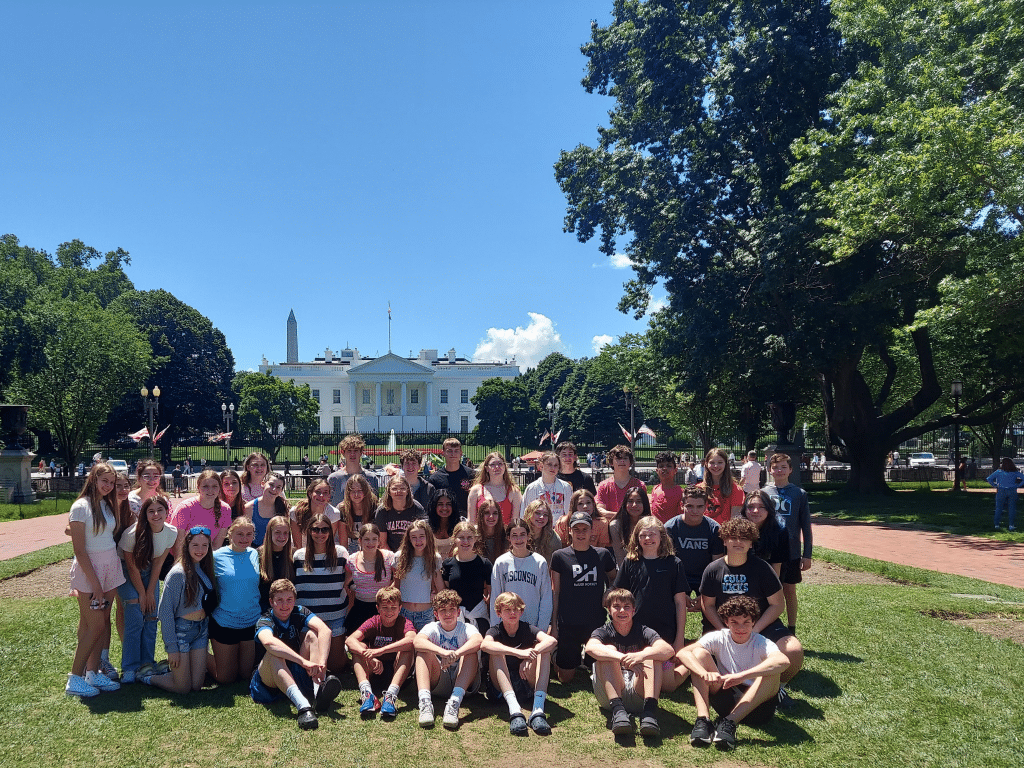 A large group of teens and a few adults pose for a photo on a green lawn with the White House and the Washington Monument behind them, enjoying a sunny, blue sky during their Landmark Educational Tours Registration experience.