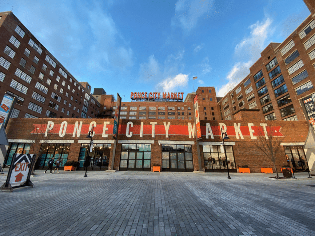 A wide-angle view of Ponce City Market, a landmark with a prominent red and white sign under a blue sky. The foreground features a paved plaza with benches, a construction sign, and banners for Landmark Educational Tours Registration.