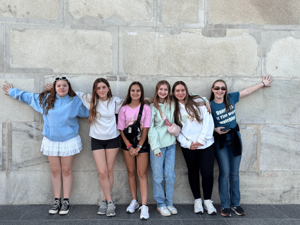 Six girls stand side by side against a large stone wall, smiling at the camera during their Landmark Educational Tours Registration. They are dressed casually in skirts, shorts, and jeans, with some wearing sunglasses or holding bags. Two girls have their arms stretched out.