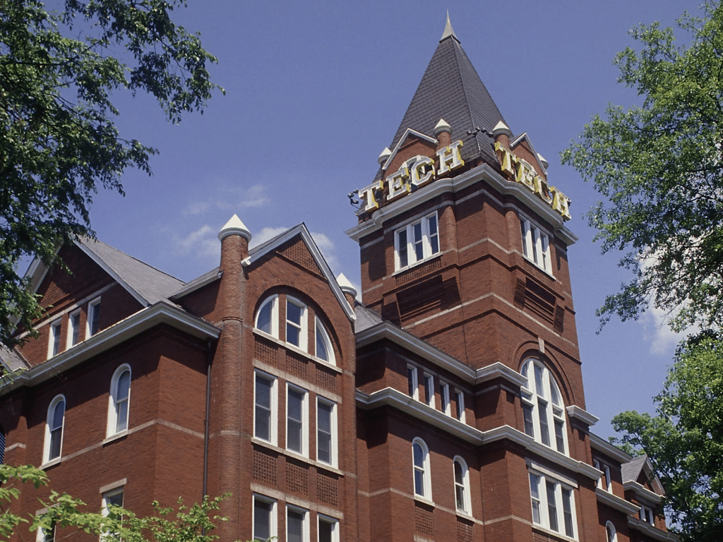 A historic red brick building with arched windows and a tall clock tower displaying "TECH" in large yellow letters—an iconic landmark perfect for Educational Tours Registration, surrounded by green trees under a clear blue sky.