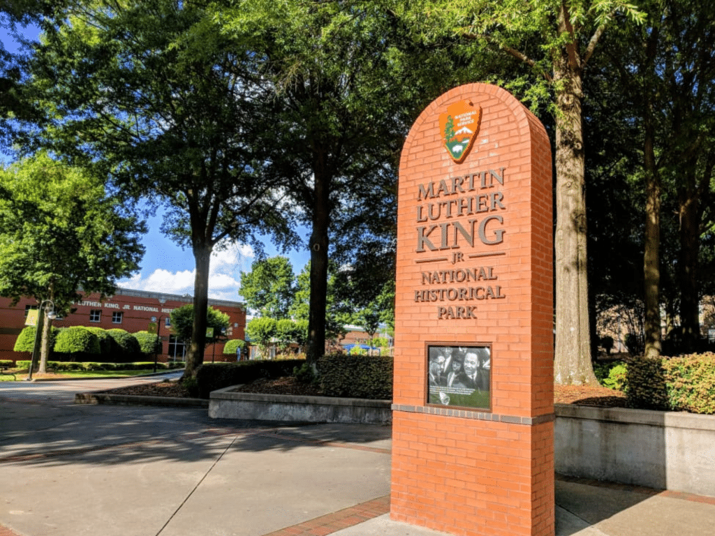 A red brick monument sign for Martin Luther King Jr. National Historical Park stands near trees and walkways, serving as a landmark for Educational Tours Registration, with a photo of Dr. King and a building in the background.