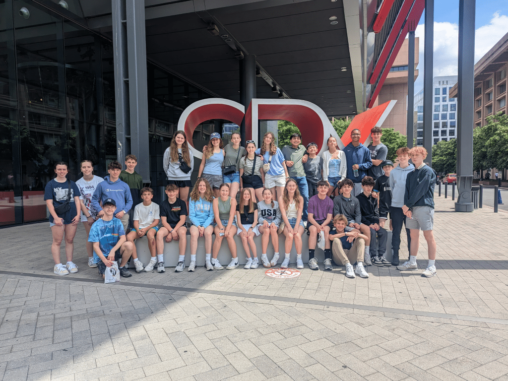 A group of about 27 teenagers pose together outside in front of a large red and white "CPX" sculpture, capturing a fun moment during their Landmark Educational Tours Registration, with a glass building and city street behind them.