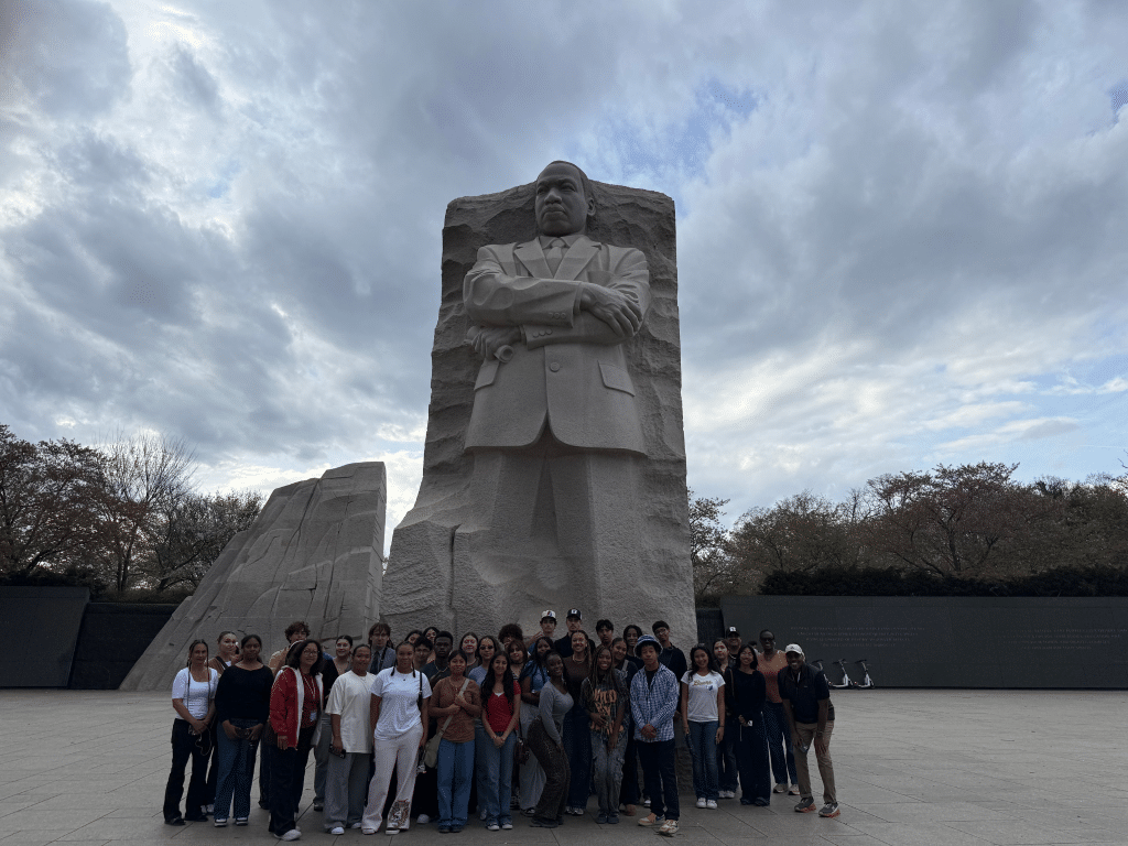 A large group of people pose for a photo in front of the Martin Luther King Jr. Memorial, a tall stone sculpture, under a cloudy sky. Trees and scooters are visible in the background.