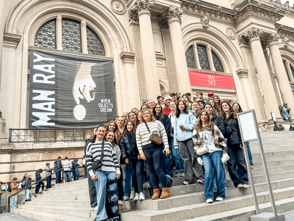 A large group poses together on the steps outside The Met museum, captured during a Landmark Educational Tours Registration. Behind them, banners read “MAN RAY: WHEN OBJECTS DREAM” and “THE MET,” framed by grand columns and arched windows.