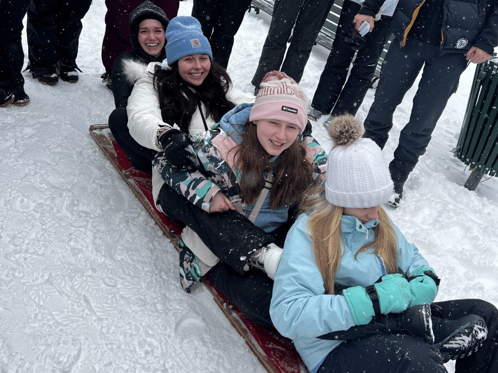 Four girls bundled in winter clothing sit on a sled in the snow, smiling and posing for a photo, while other people stand in the background. Snow is falling and everyone appears to be enjoying a winter day.